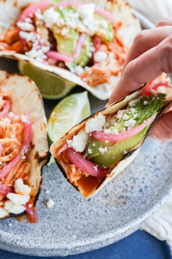 A hand holds a taco filled with shredded chicken, avocado, pickled red onions, and crumbled cheese, with lime wedges and more tacos on a speckled plate in the background.