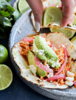 A hand squeezes a lime wedge over chicken tinga tacos filled with shredded chicken, avocado slices, pickled onions, and crumbled cheese on a grilled tortilla. Fresh limes and cilantro are nearby on a blue plate.