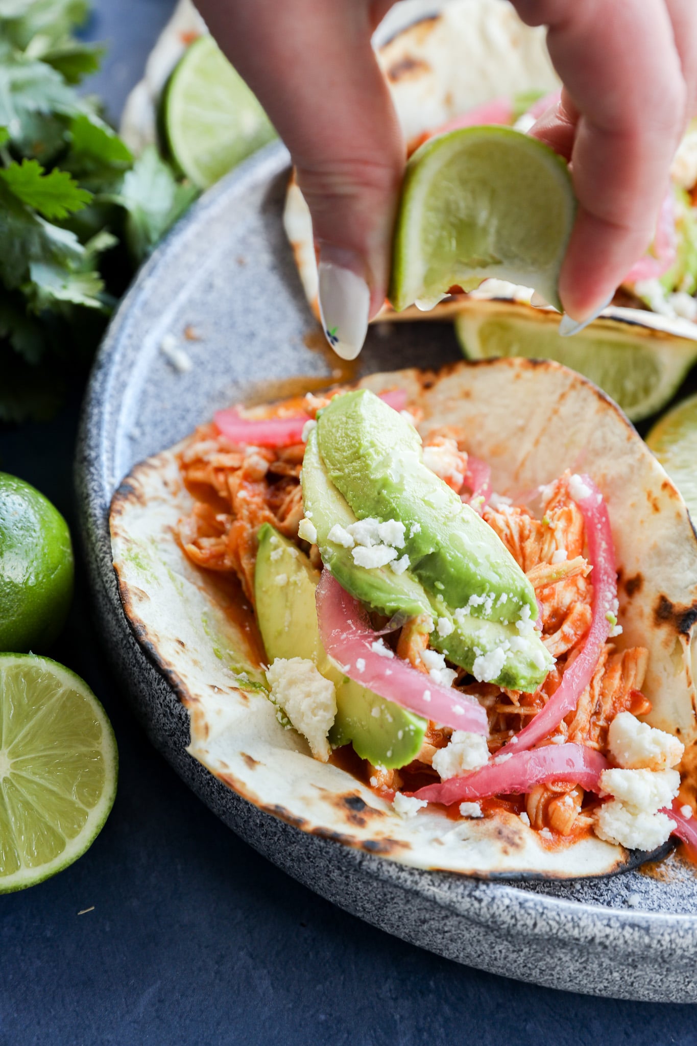 A hand squeezes a lime wedge over chicken tinga tacos filled with shredded chicken, avocado slices, pickled onions, and crumbled cheese on a grilled tortilla. Fresh limes and cilantro are nearby on a blue plate.