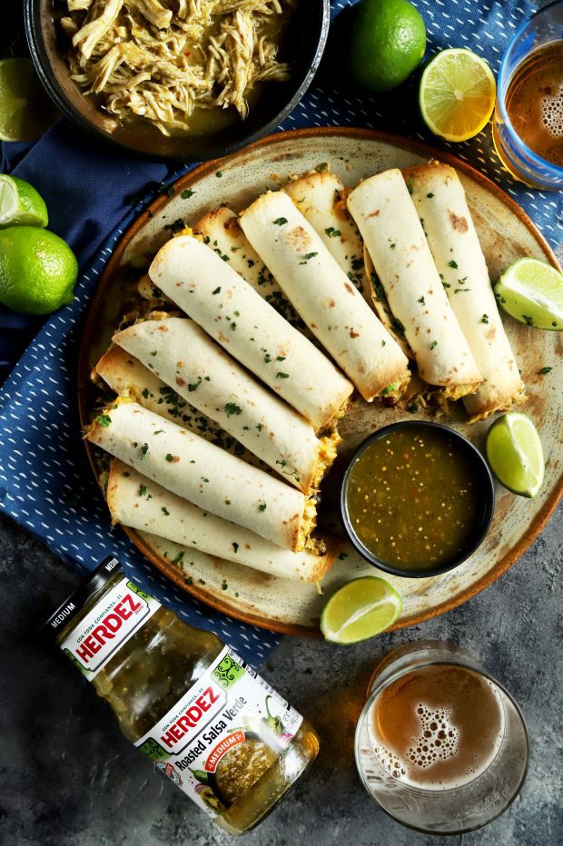 A pile of baked taquitos for a party with a beer and jar of salsa verde