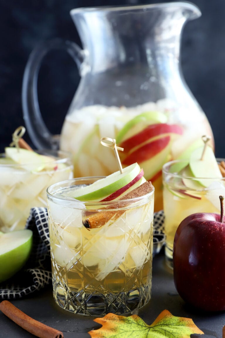 apple pie sparkling sangria in glasses with apples and cinnamon sticks, a pitcher of the cocktail in the background