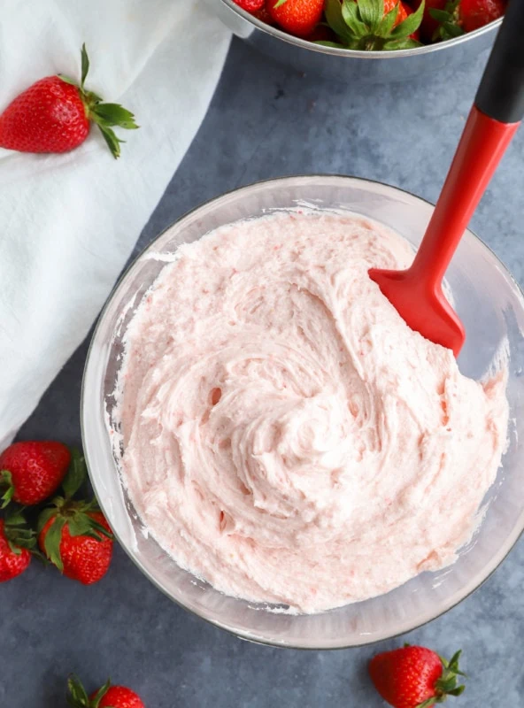 Strawberry buttercream frosting in a bowl with a spatula and fresh fruit