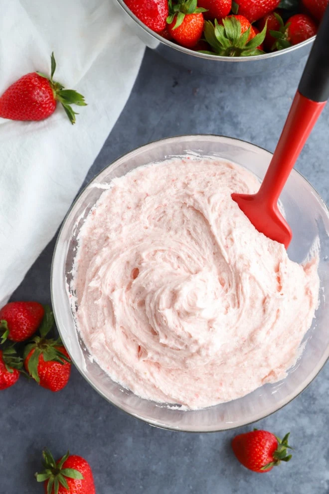 Strawberry buttercream frosting in a bowl with a spatula and fresh fruit