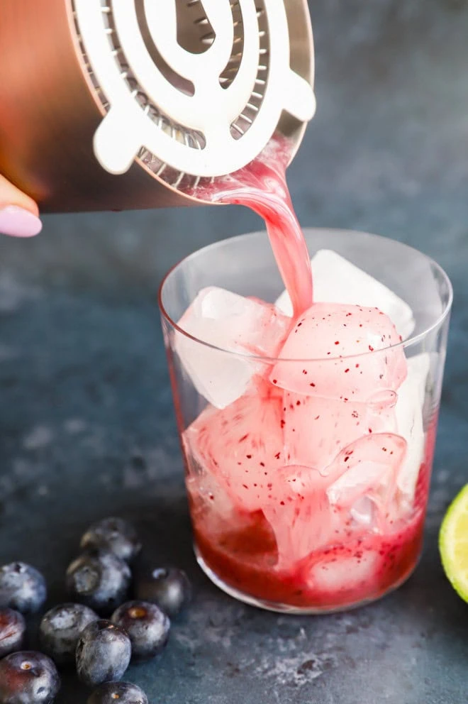 straining blueberry margarita into a glass filled with ice