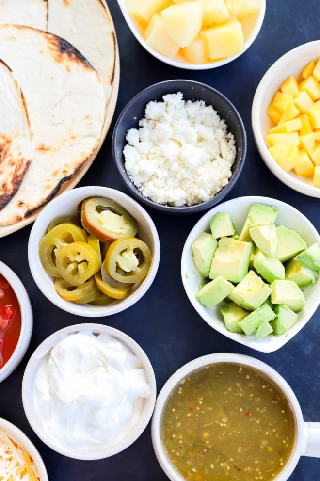 Bowls of chopped mango, pineapple, avocado, sour cream, salsa verde, and pickled jalapeno slices and corn tortillas