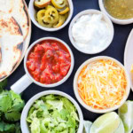 Image of a variety of toppings including shredded lettuce, lime wedges, shredded cheese, sour cream, salsa verde, salsa, pickled jalapeno, and tortillas
