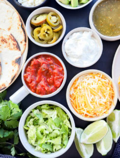 Image of a variety of toppings including shredded lettuce, lime wedges, shredded cheese, sour cream, salsa verde, salsa, pickled jalapeno, and tortillas