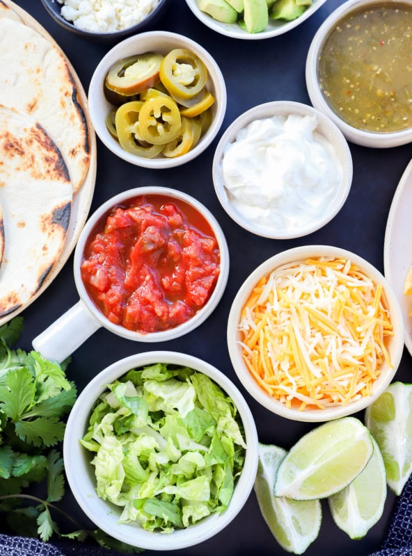 Image of a variety of toppings including shredded lettuce, lime wedges, shredded cheese, sour cream, salsa verde, salsa, pickled jalapeno, and tortillas