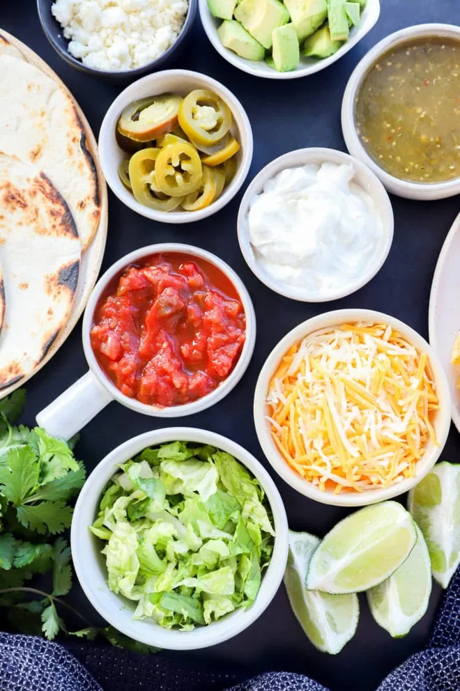 Image of a variety of toppings including shredded lettuce, lime wedges, shredded cheese, sour cream, salsa verde, salsa, pickled jalapeno, and tortillas