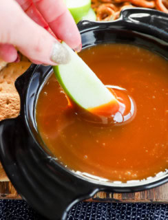 hand dipping an apple slice in caramel apple dip in a bowl