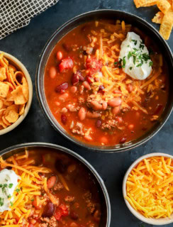 bowls of instant pot chili topped with cheese, sour cream, and parsley with fritos on the side