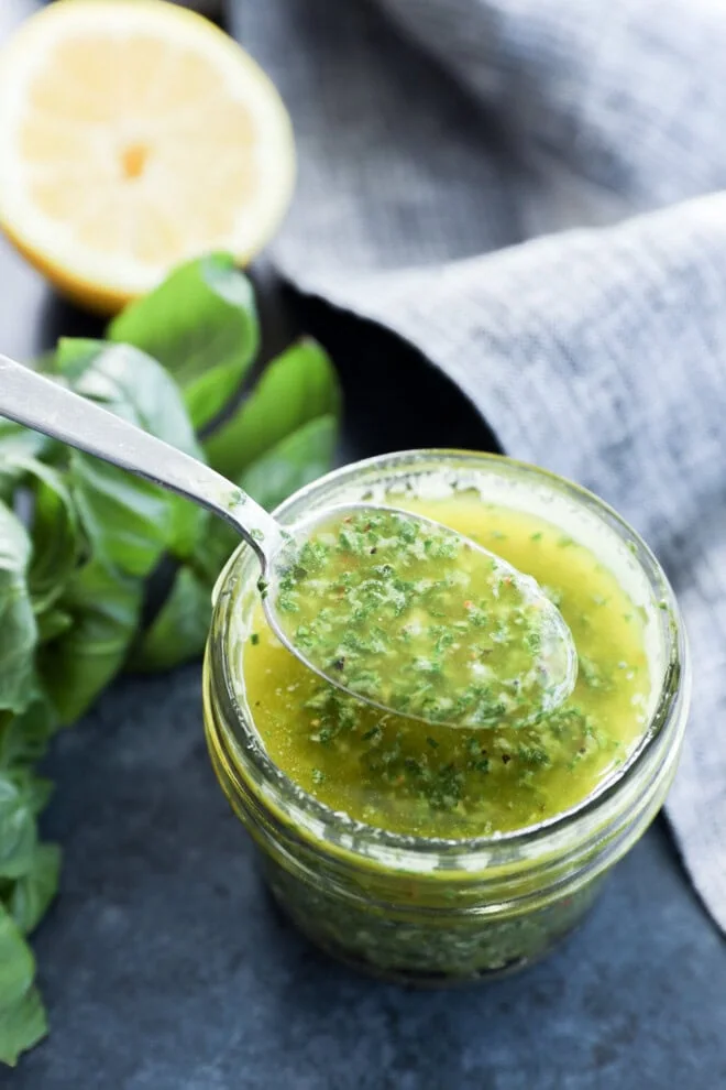Spooning basil oil out of a mason jar with fresh basil and lemon in the background