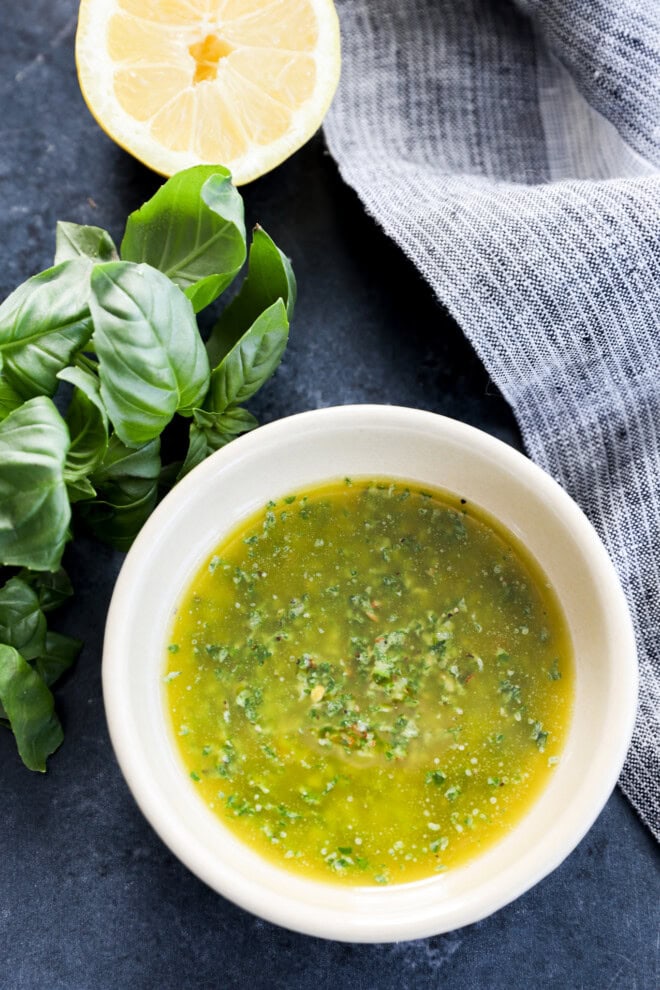 Image of basil oil in a bowl with fresh basil sprigs and half a lemon on a grey linen