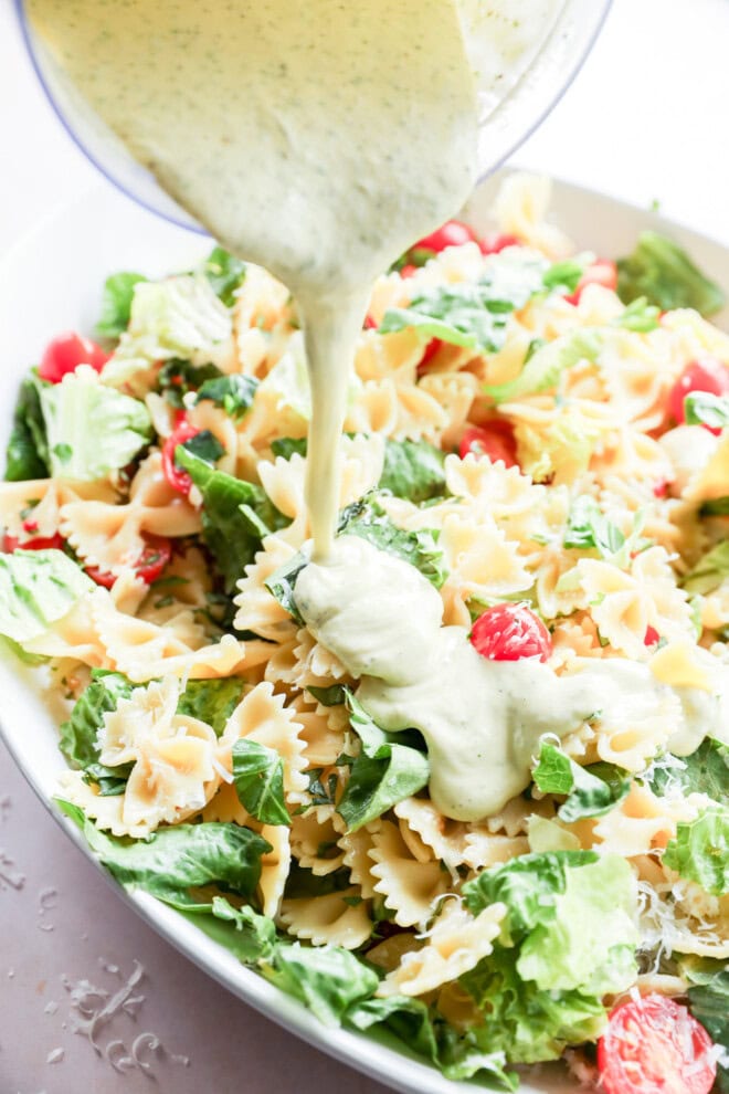 pouring basil dressing on top of a side dish of lettuce, penne, and vegetables