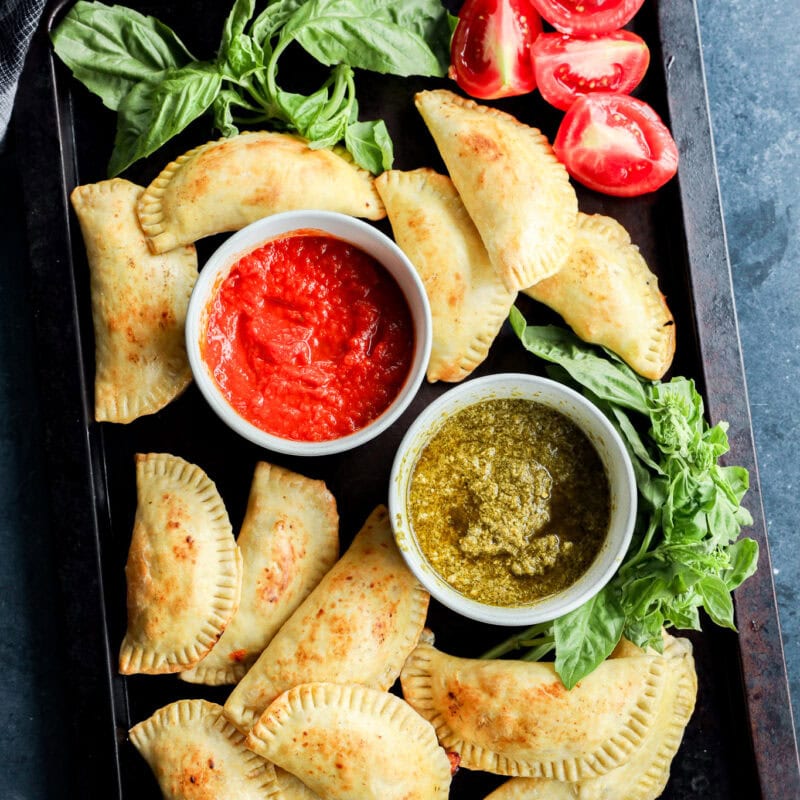 caprese empanadas on sheet pan with dipping sauces, fresh basil, and tomatoes