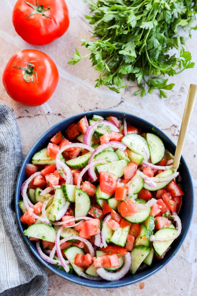 Image of Cucumber tomato salad in a bowl with gold spoon, tomatoes and parsley in the background
