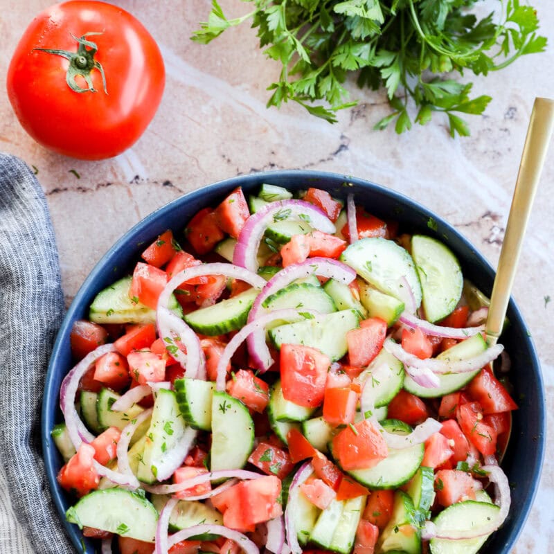 Image of Cucumber tomato salad in a bowl with gold spoon, tomatoes and parsley in the background