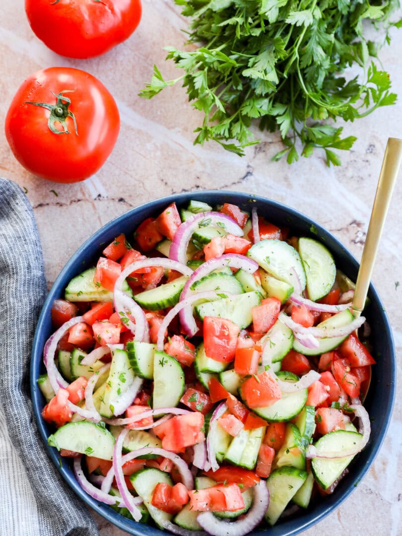 Image of Cucumber tomato salad in a bowl with gold spoon, tomatoes and parsley in the background