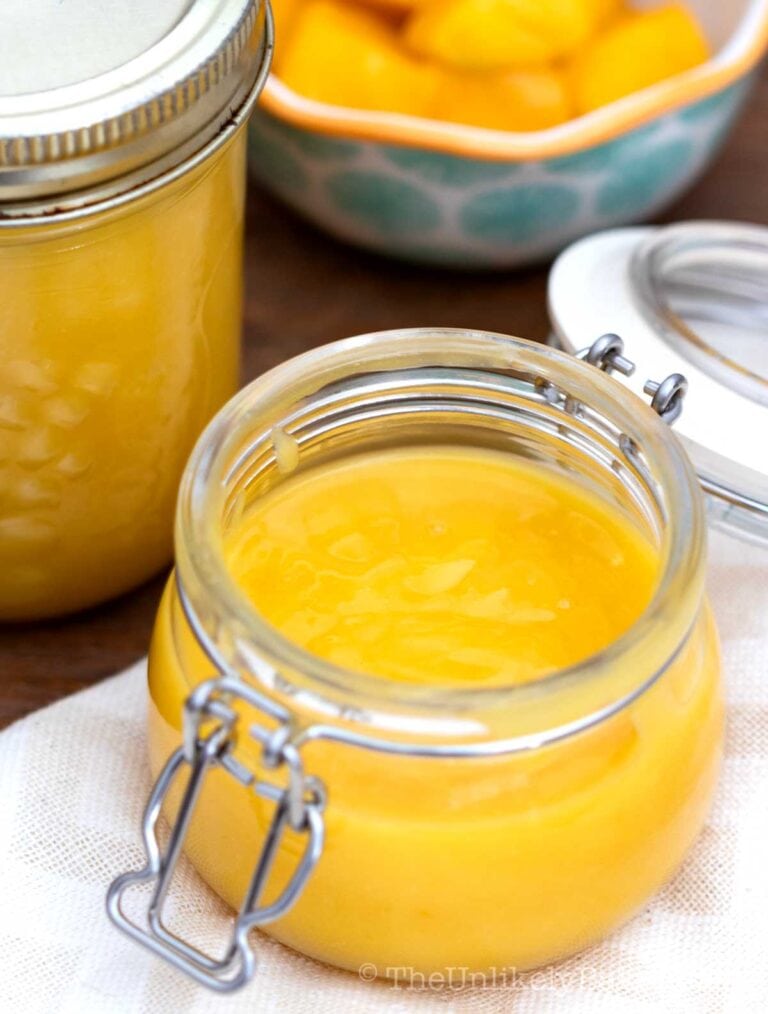 An open glass jar filled with smooth yellow lemon curd sits on a white cloth, with a sealed jar and a bowl of chopped mango in the background.