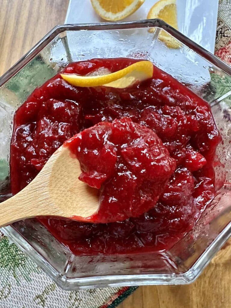 A glass bowl filled with thick cranberry sauce, being stirred with a wooden spoon. A lemon peel garnish is on top, and orange slices are visible in the background.