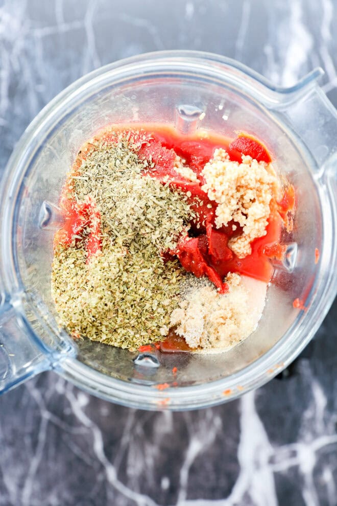 A blender filled with tomato paste, minced garlic, dried oregano, sugar, and other spices—the perfect blend for the Best Pizza Sauce—viewed from above on a marbled countertop.