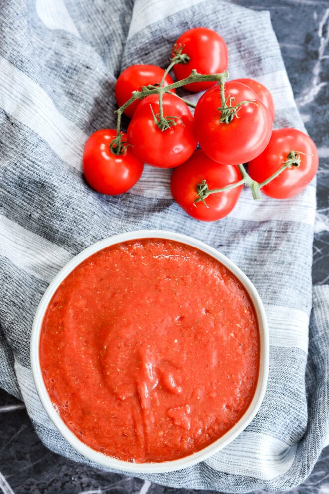 A cluster of fresh tomatoes on the vine sits next to a bowl of smooth tomato sauce arranged on a striped gray and white cloth atop a dark marble surface.