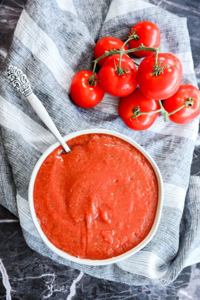 A bowl of smooth red tomato sauce, perfect as the best pizza sauce, with a spoon rests on a striped cloth beside fresh tomatoes on the vine, all set on a dark marble surface.