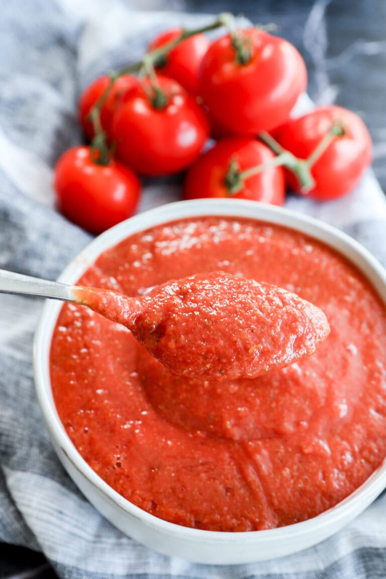 A spoonful of thick, Best Pizza Sauce is held above a bowl filled with more sauce, with fresh vine tomatoes in the background on a striped cloth.