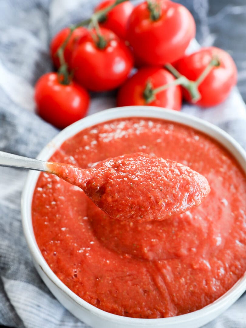 A spoonful of thick, Best Pizza Sauce is held above a bowl filled with more sauce, with fresh vine tomatoes in the background on a striped cloth.