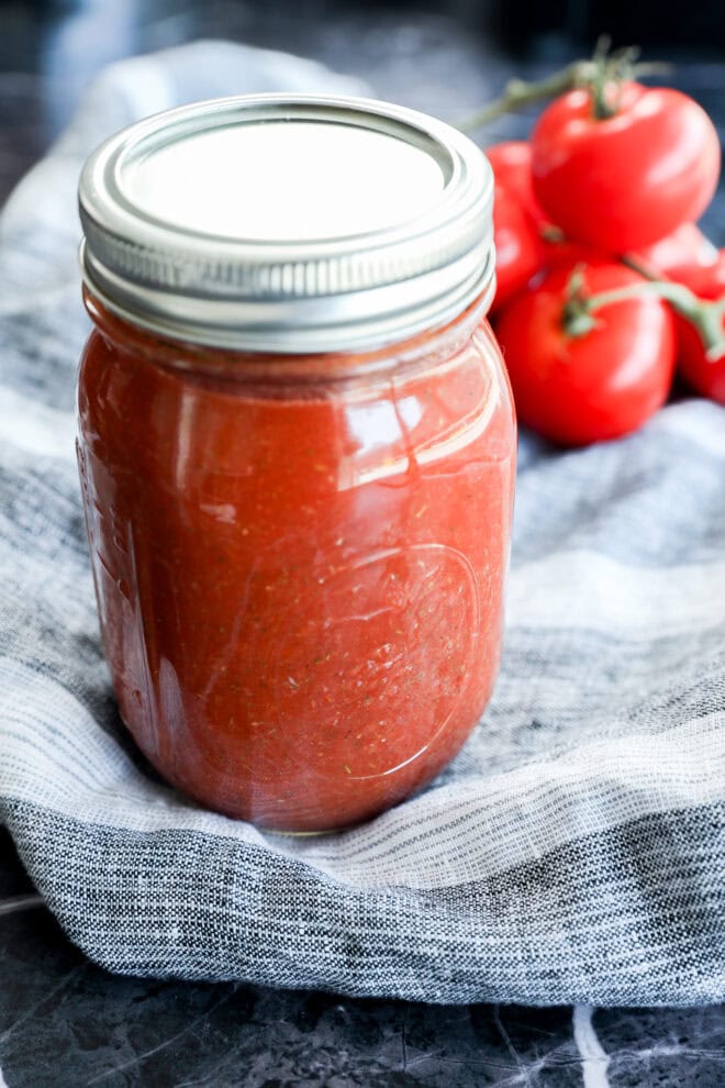 A glass mason jar filled with red tomato sauce, known as the best pizza sauce, sits on a striped cloth, with fresh tomatoes on the vine in the background.