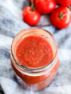 A glass jar filled with red tomato sauce, perfect as the best pizza sauce, sits on a striped cloth, with several fresh tomatoes on the vine in the background.