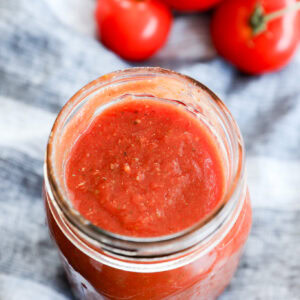 A glass jar filled with red tomato sauce, perfect as the best pizza sauce, sits on a striped cloth, with several fresh tomatoes on the vine in the background.
