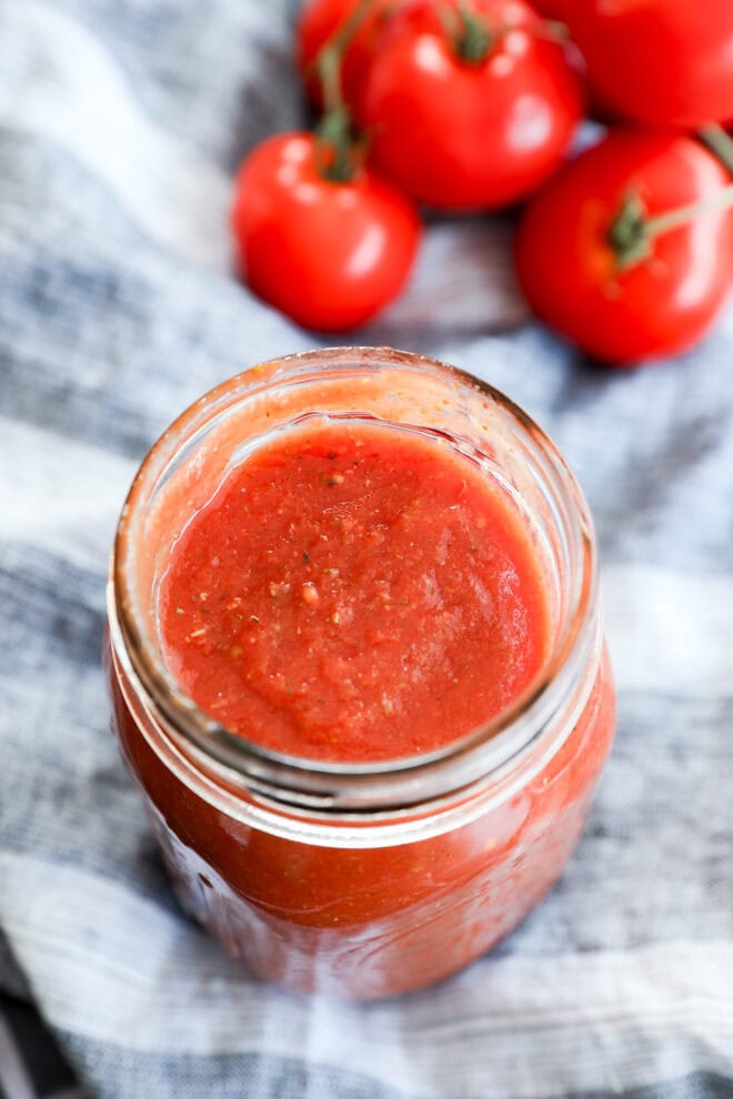 A glass jar filled with red tomato sauce, perfect as the best pizza sauce, sits on a striped cloth, with several fresh tomatoes on the vine in the background.
