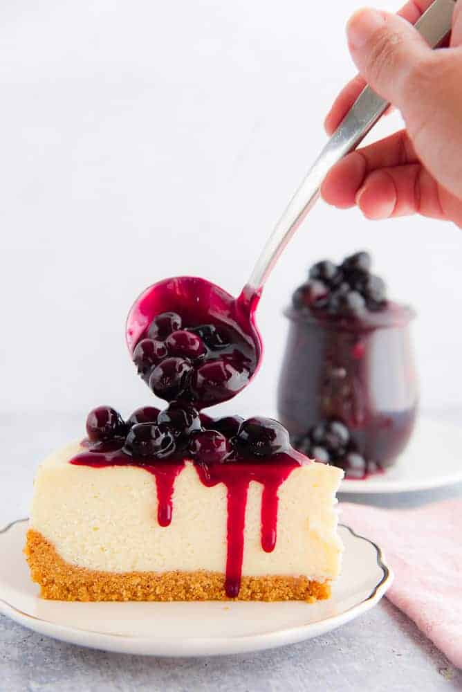 A hand spooning blueberry sauce onto a slice of cheesecake with a graham cracker crust, on a white plate. A jar of blueberry sauce is blurred in the background.