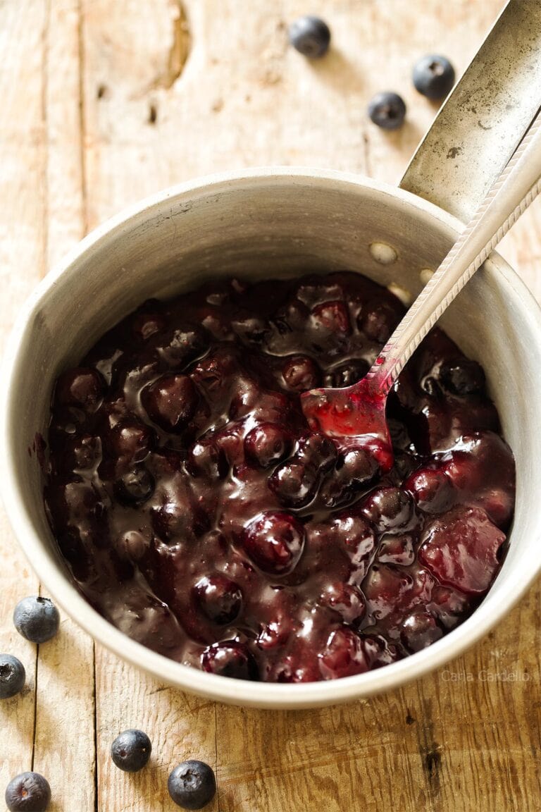 A metal saucepan filled with thick, glossy blueberry sauce, with whole blueberries visible. A spoon rests inside the pan, and a few fresh blueberries are scattered on the wooden surface nearby.