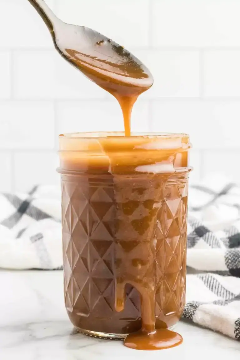 A glass jar filled with butterscotch sauce, with sauce dripping down the side. A spoon above the jar is drizzling more caramel into it. A towel with a checkered pattern is in the background.