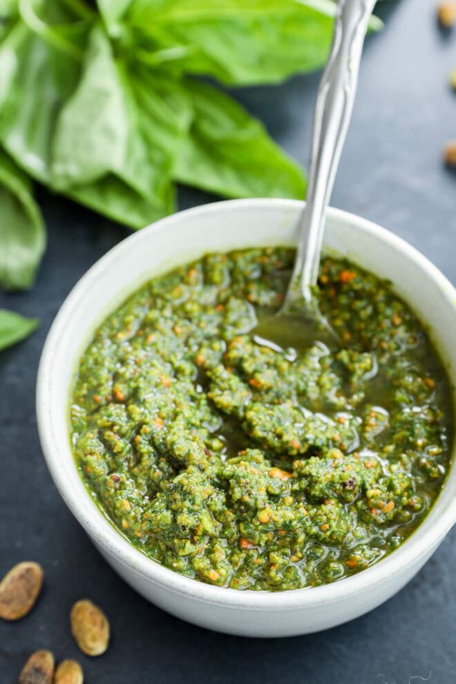 White bowl full of basil sauce with spoon in it, nuts on the side and fresh basil in the background