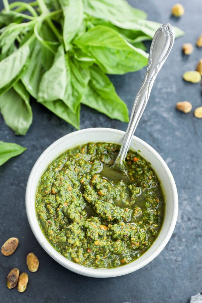 Basil sauce in white bowl with spoon in it next to a bunch of fresh basil