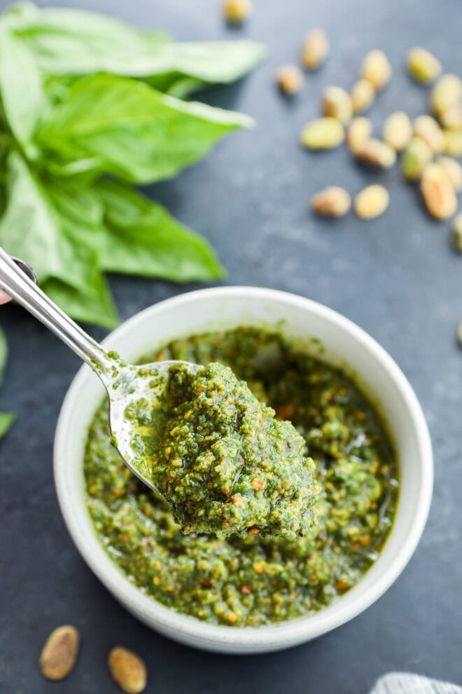 Spoon of pistachio pesto lifted out of the white bowl with nuts on the side and a bunch of fresh basil