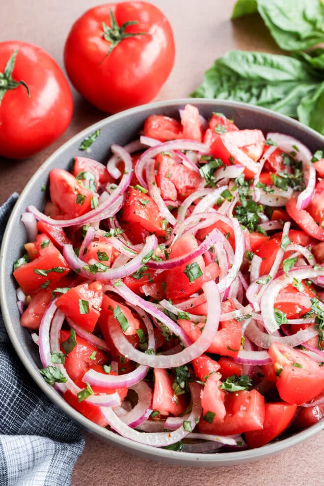 Tomato onion salad in a bowl with tomatoes and fresh basil behind it