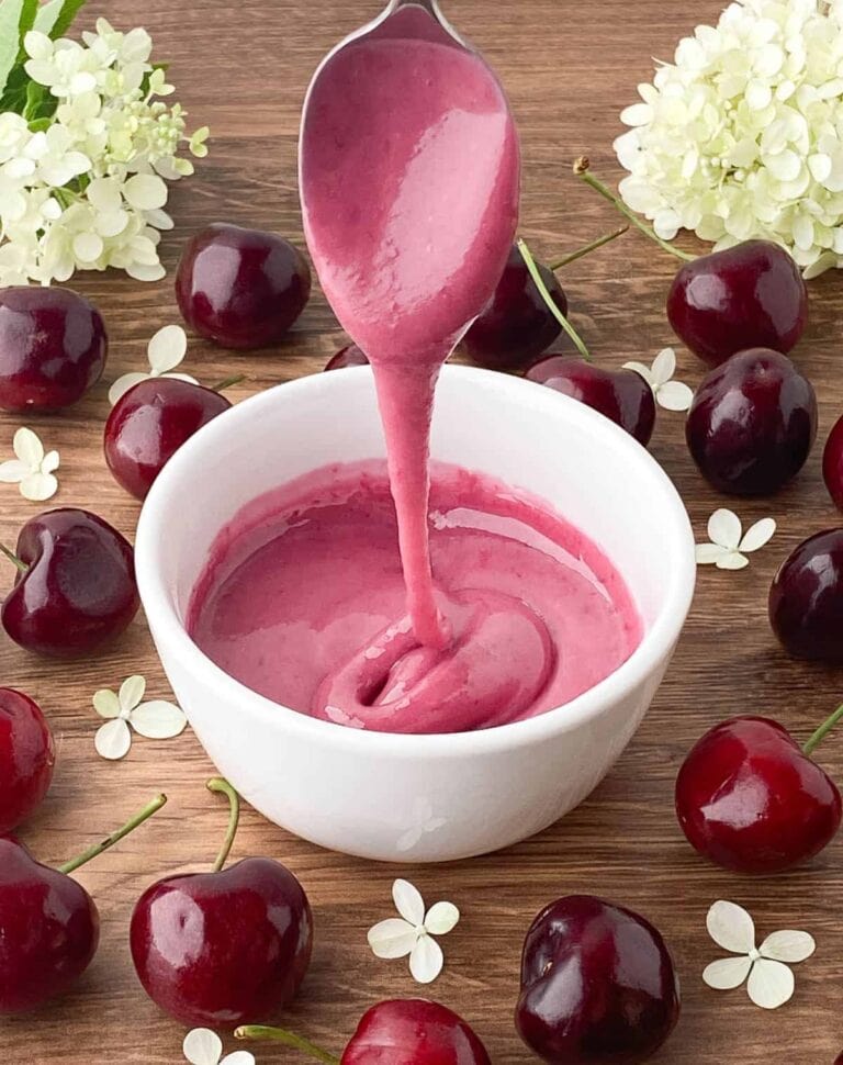 A spoon drizzles thick, pink cherry sauce into a white bowl, surrounded by fresh cherries and white flowers on a wooden surface.