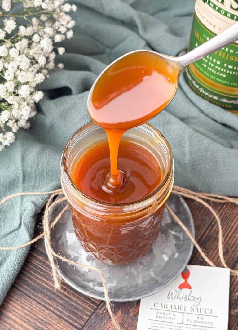 A spoon drizzles thick caramel sauce into a glass jar. The jar is tied with twine and sits on a plate next to a label reading “Whiskey Caramel Sauce.” A bottle and white flowers are in the background.