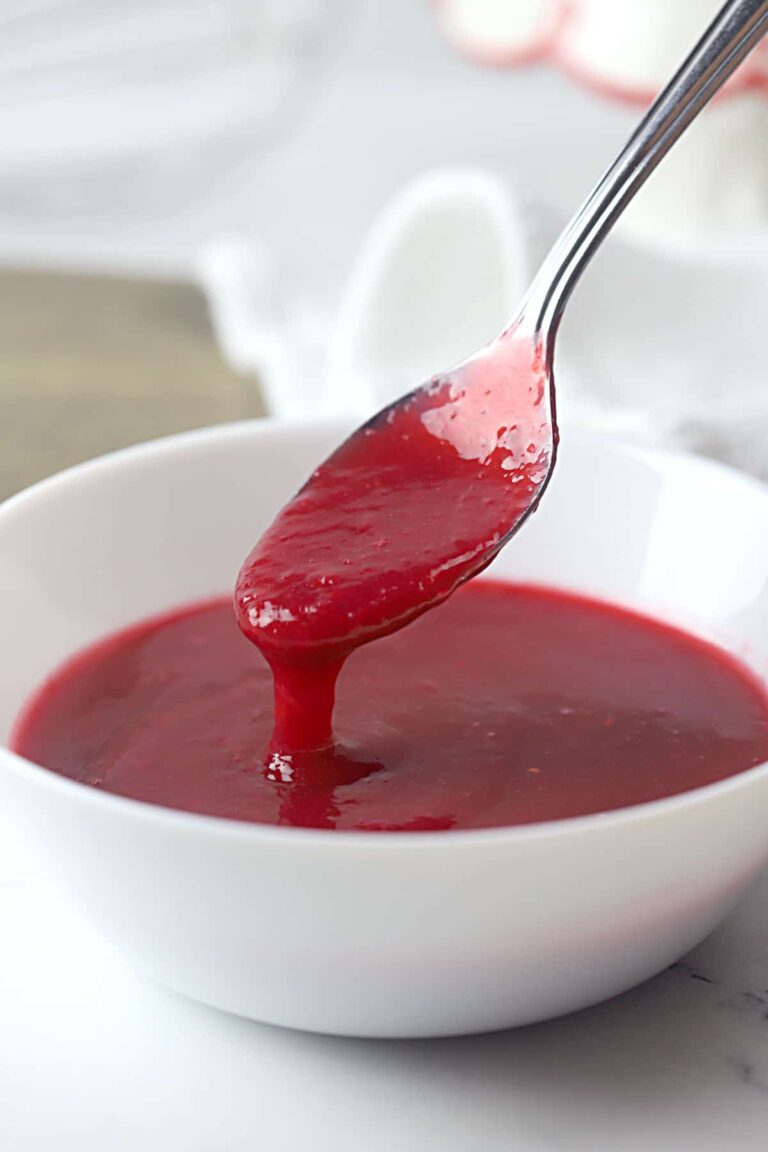 A spoon holding up a thick, glossy red sauce or coulis above a white bowl filled with the same sauce, set on a white surface.