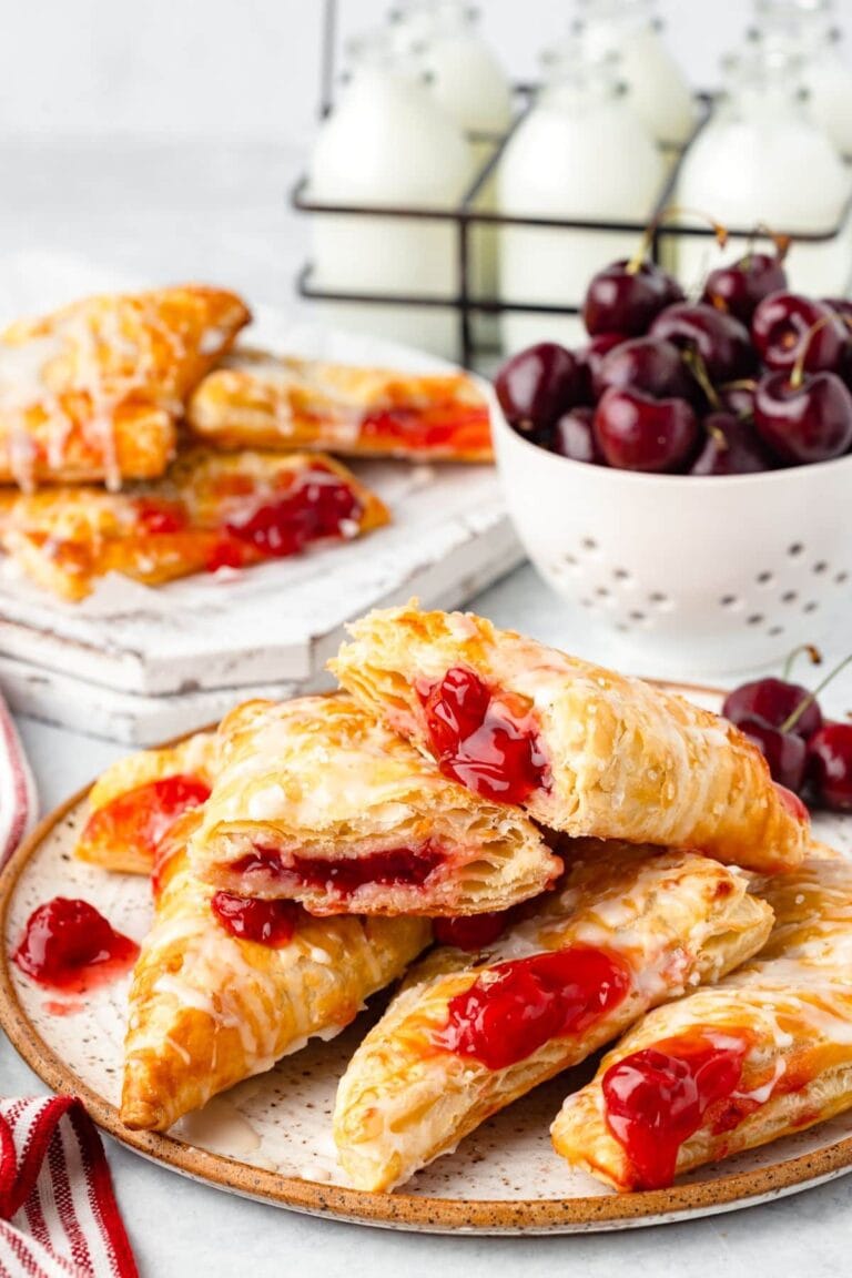 Golden, flaky cherry turnovers—classic puff pastry dessert recipes—are stacked on a plate and cutting board. A bowl of fresh cherries and glass bottles of milk in a metal carrier are in the background.