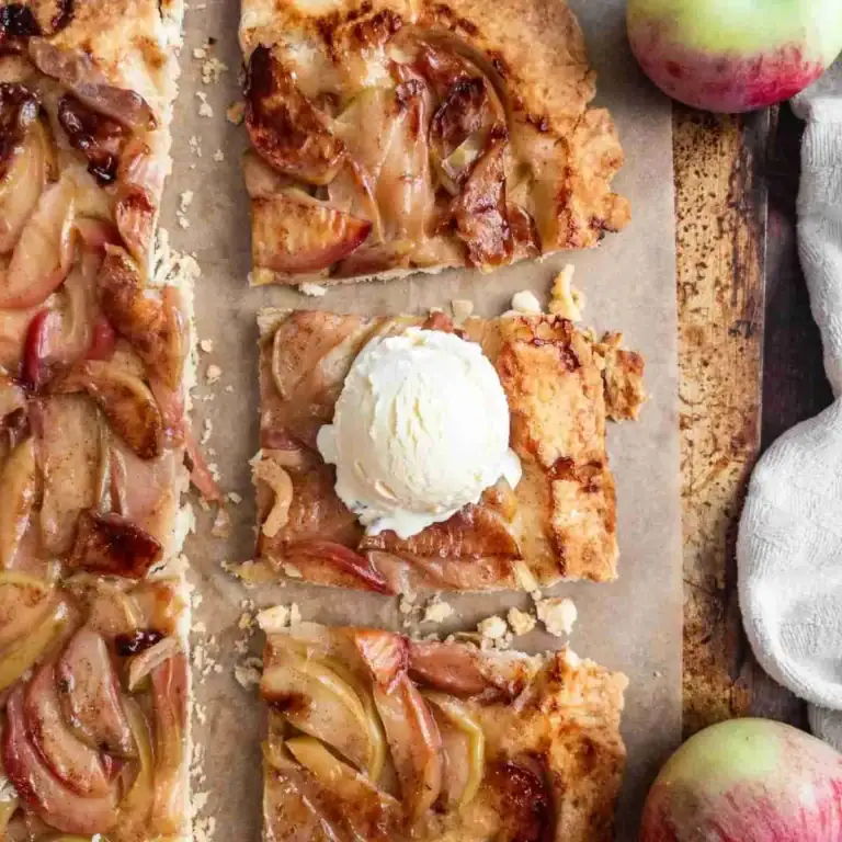 Rectangular slices of apple tart made with puff pastry dessert recipes rest on parchment paper, one topped with vanilla ice cream. Whole apples and a white cloth sit nearby on a rustic surface.