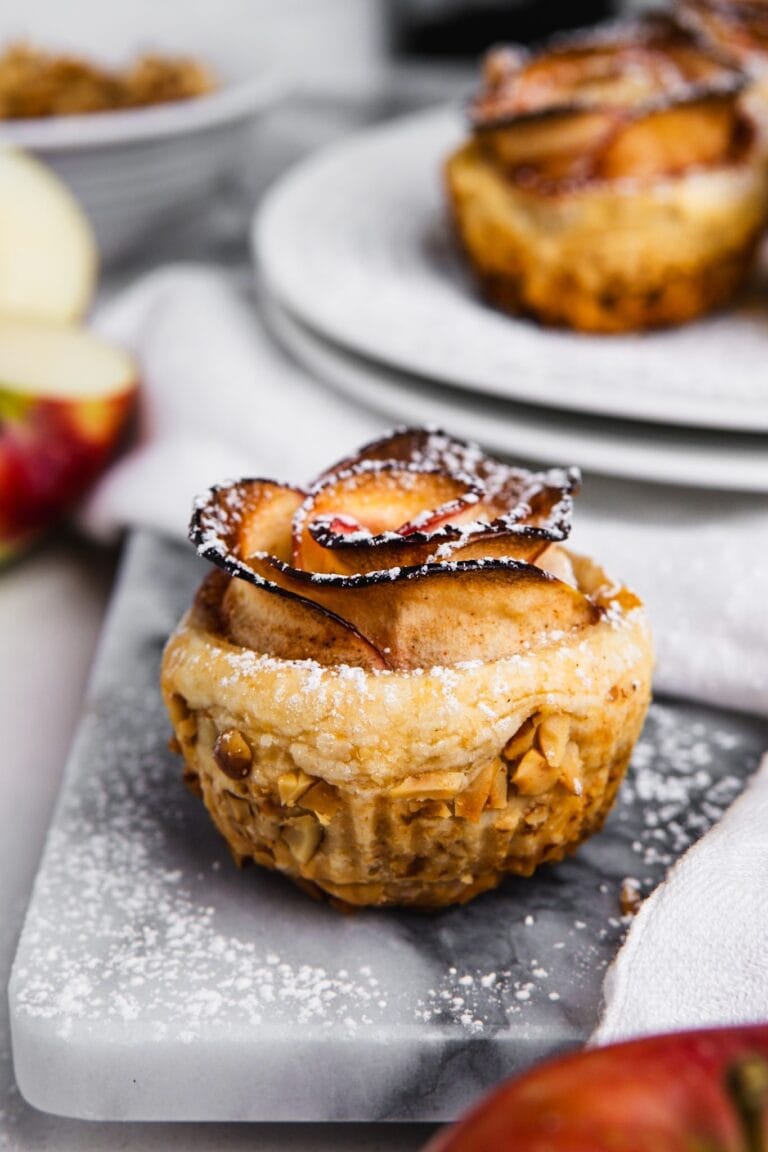A close-up of a puff pastry dessert, topped with thinly sliced apples arranged like a rose and dusted with powdered sugar, sits on a marble surface with blurred pastries and apples in the background—perfect for puff pastry dessert recipes.