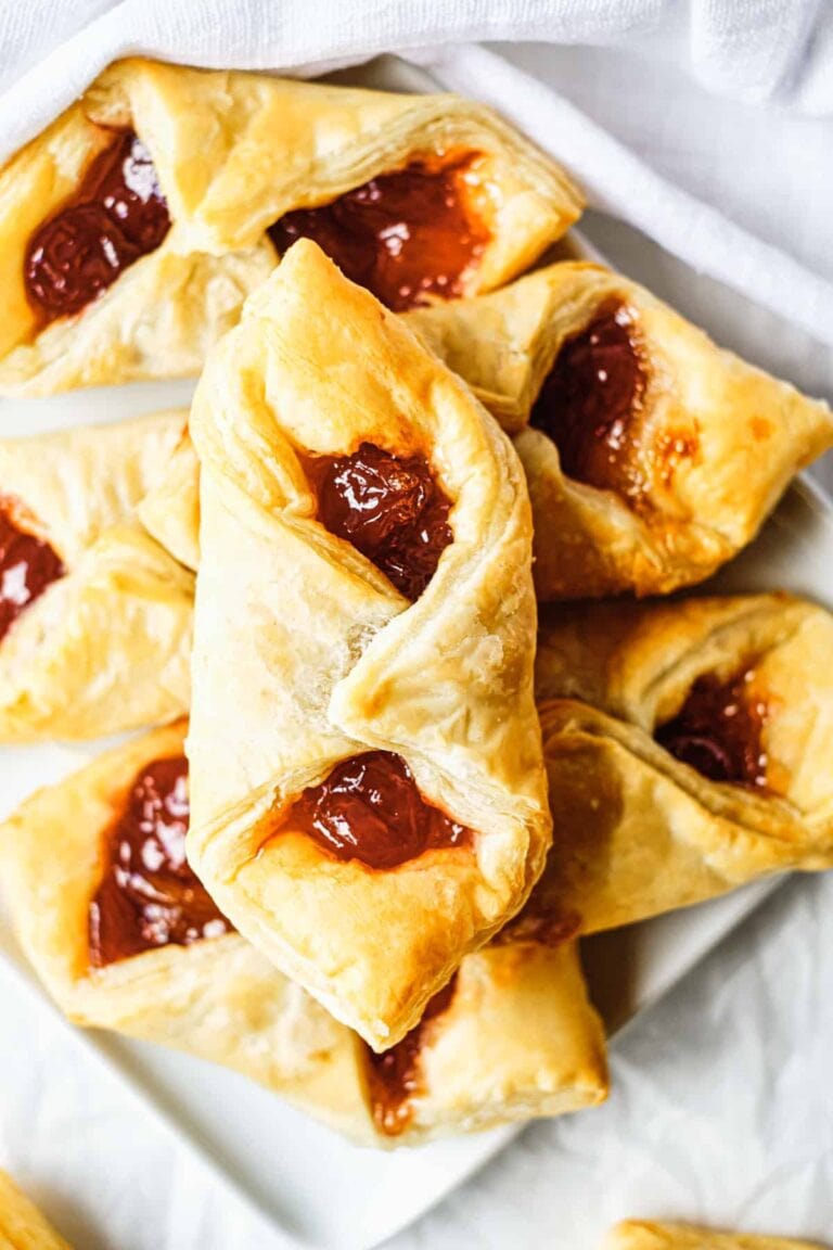 A plate of golden, flaky pastries filled with bright red fruit jam, arranged in a slightly overlapping pile on a white surface—perfect inspiration for your next puff pastry dessert recipes.