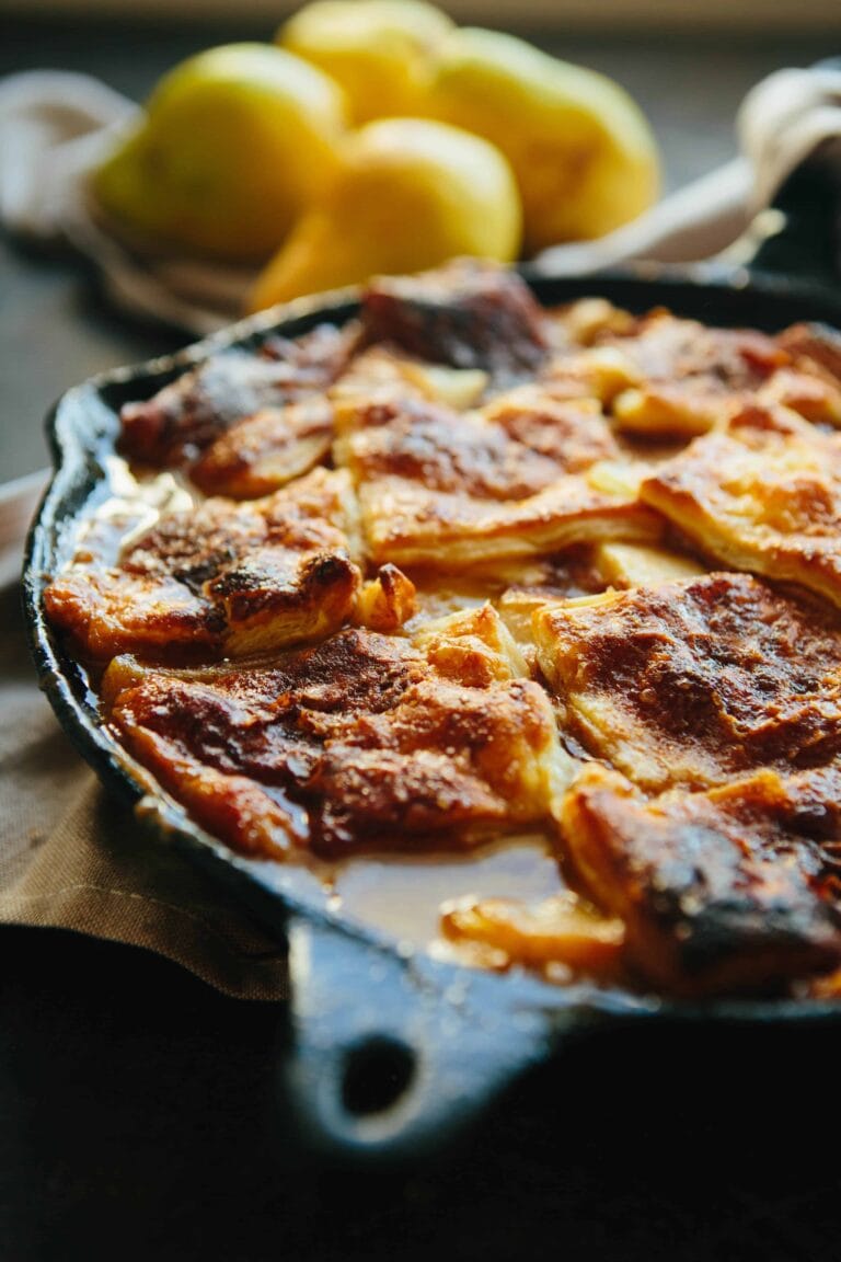 A close-up of a golden-brown baked dessert in a cast iron skillet, with slightly crispy edges and creamy filling—perfect inspiration for puff pastry dessert recipes. Out-of-focus lemons are in the background.
