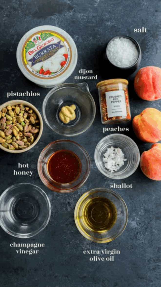 Top-down view of ingredients on a dark surface: burrata cheese, pistachios, hot honey, Dijon mustard, champagne vinegar, extra virgin olive oil, salt, crushed pepper, shallot, and two fresh peaches.
