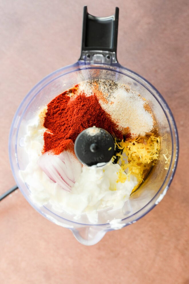 A food processor bowl containing yogurt, paprika, garlic powder, lemon zest, chopped shallot, and olive oil, viewed from above on a light brown surface.